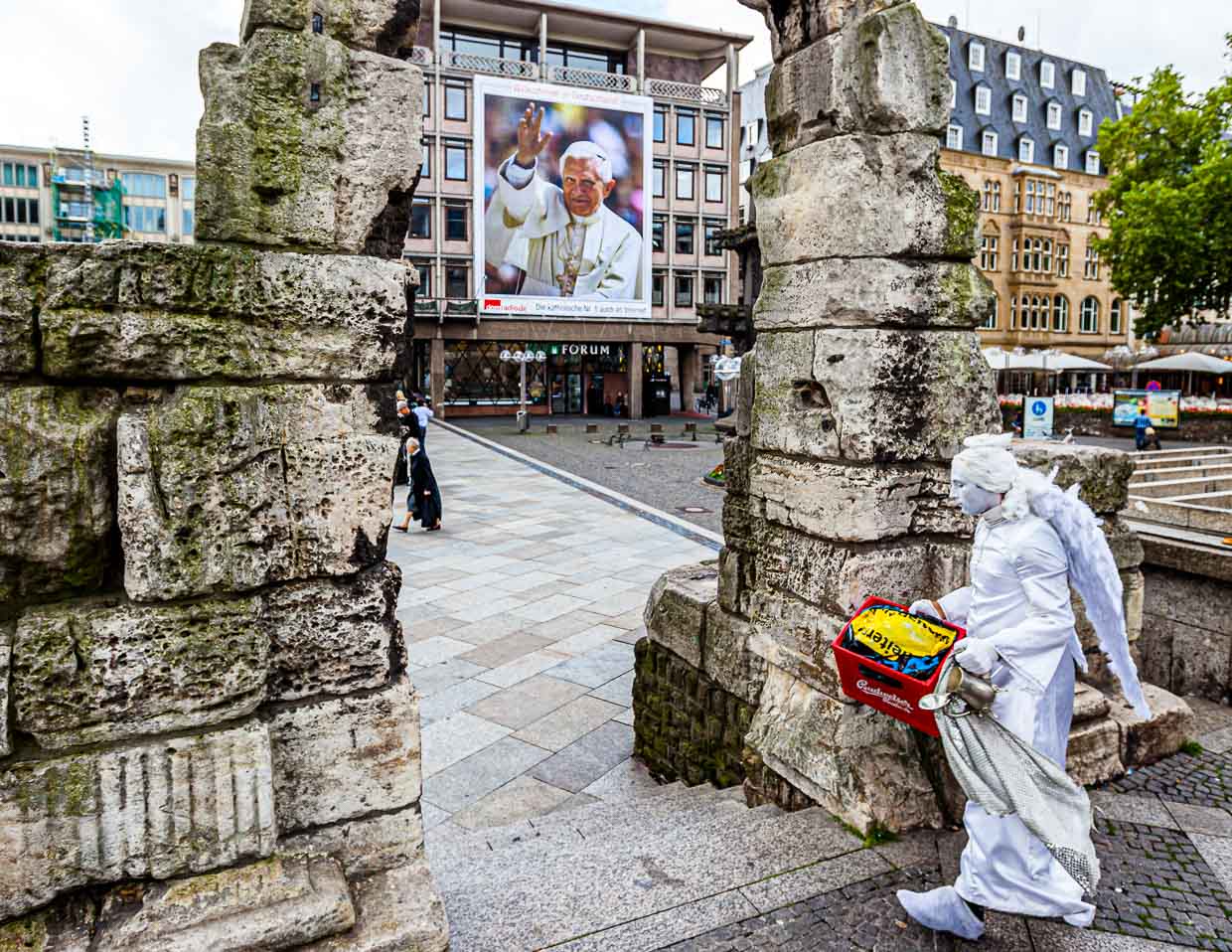 Angel with beer crate in front of the Pope. A living statue strides through the Roman Gate in front of Cologne Cathedral with a beer crate, which it will use as a pedestal. In the background Pope Benedict VI waves in survival size from a poster / © Photo: Georg Berg